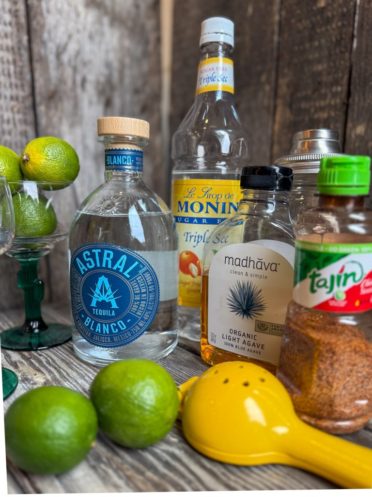 Skinny margarita ingredients on a rustic wood surface including fresh limes, Astral tequila, Monin sugar free triple sec syrup, Madhava organic agave, Tajín seasoning, and margarita glasses.
