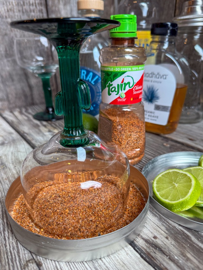 Glass margarita cup with cactus stem being rimmed in Tajín seasoning beside sliced limes and cocktail ingredients on a wooden table.