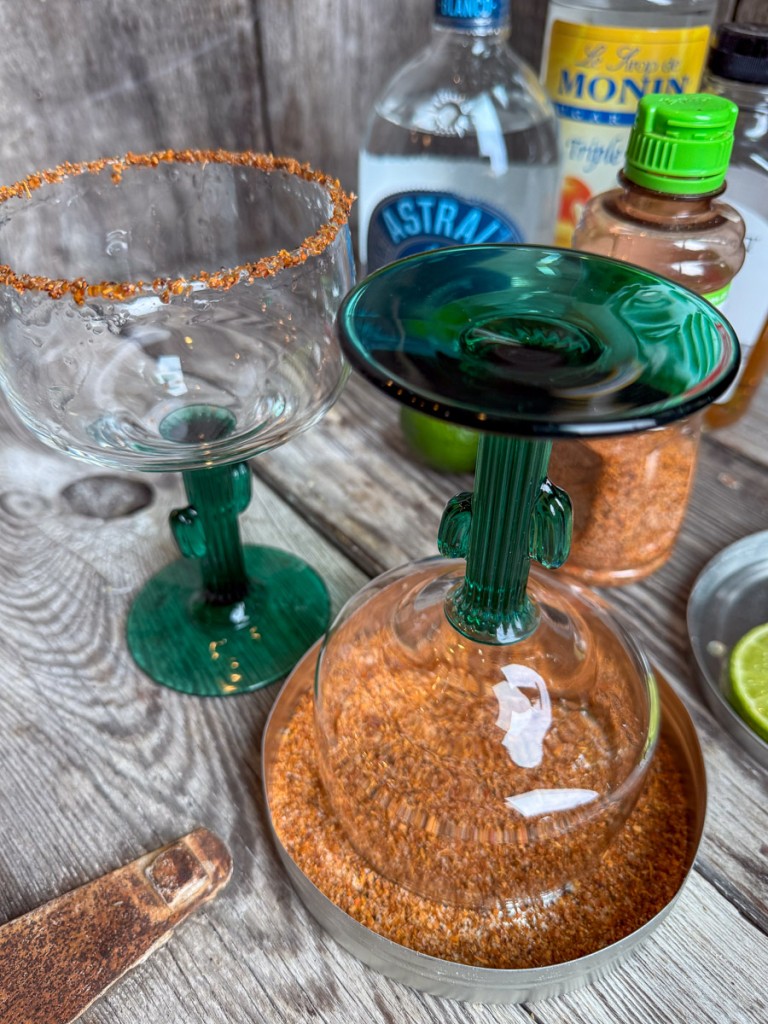 Glass margarita cup with cactus stem being rimmed in Tajín seasoning beside sliced limes and cocktail ingredients on a wooden table.