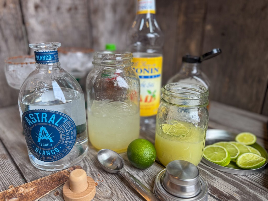 Mason jars filled with fresh lime juice and mixed margarita beside bottles of Astral tequila, sugar free triple sec syrup, and agave on a rustic surface.