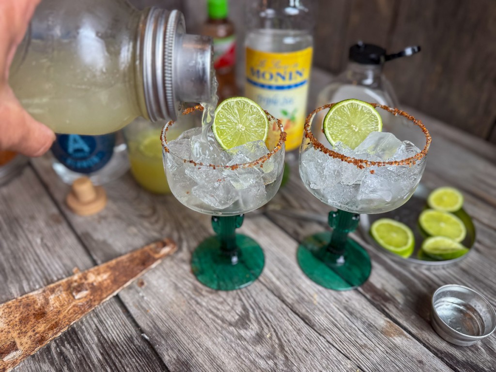 Hand pouring a freshly shaken skinny margarita into two cactus-stem margarita glasses rimmed with Tajín and filled with ice and lime slices.