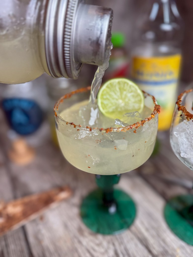Close-up pour of a skinny margarita from a mason jar shaker into a Tajín-rimmed margarita glass with a lime wedge.