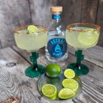 Finished skinny margaritas in cactus glasses with Tajín rims and lime slices, set next to a bottle of Astral tequila and sliced limes on a metal plate.