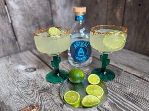 Finished skinny margaritas in cactus glasses with Taj&iacute;n rims and lime slices, set next to a bottle of Astral tequila and sliced limes on a metal plate.