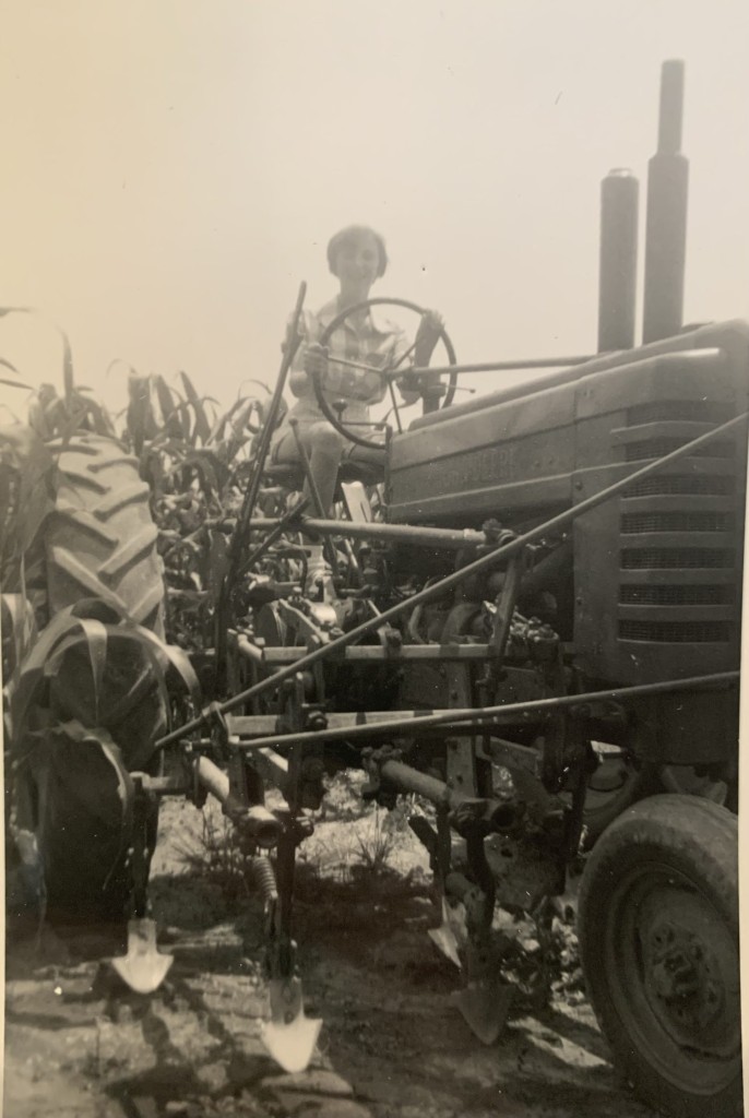 Black and white photo of a young woman on an old tractor in a cornfield, representing a lifelong farm wife's early days on the farm