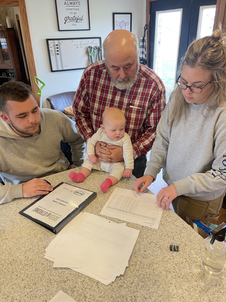 Three generations of a farming family, including an infant held by an older man in a plaid shirt, reviewing documents with a younger mand and woman at a kitchen counter. Image showcasing family involvement in farm business planning and financial management.