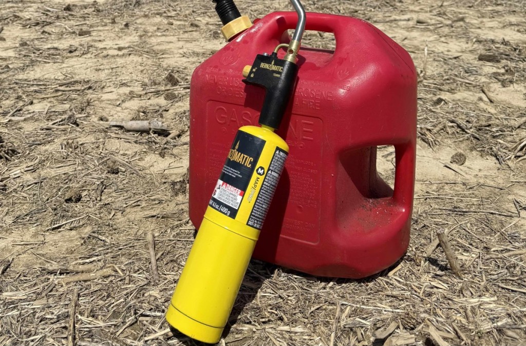 A yellow Bernzomatic blow torch leaning against a red gasoline container of dry, mulch-covered ground-used for farm maintenance or repairs.