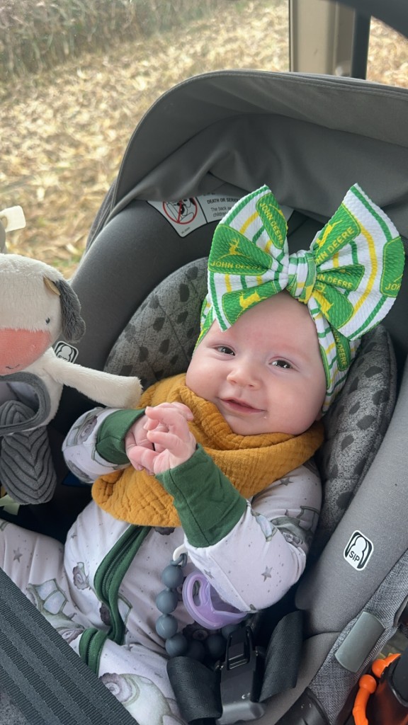 Baby girl smiling in car seat wearing tractor-print pajamas and green John Deere bow, holding cow toy