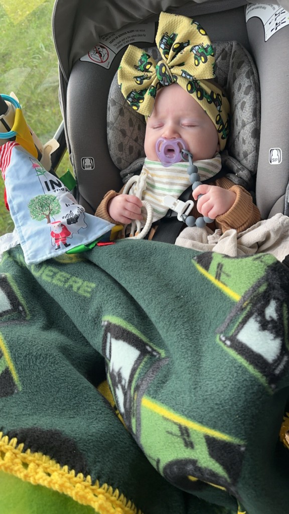 Baby girl asleep under green John Deere blanket wearing yellow tractor-print bow in tractor cab