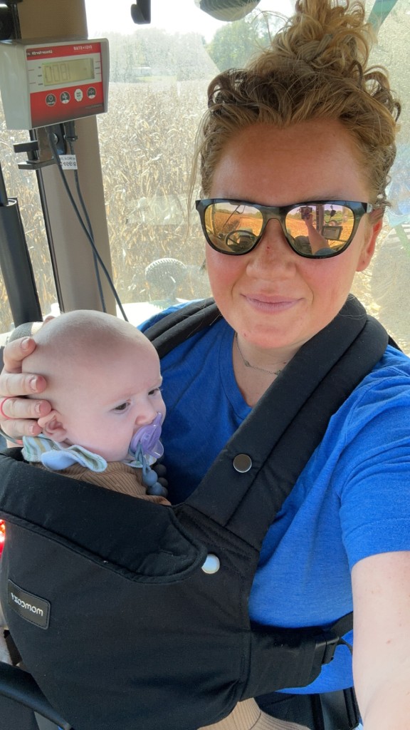 Smiling Farm mom hlding baby in a carrier inside tractor cab during corn harvest