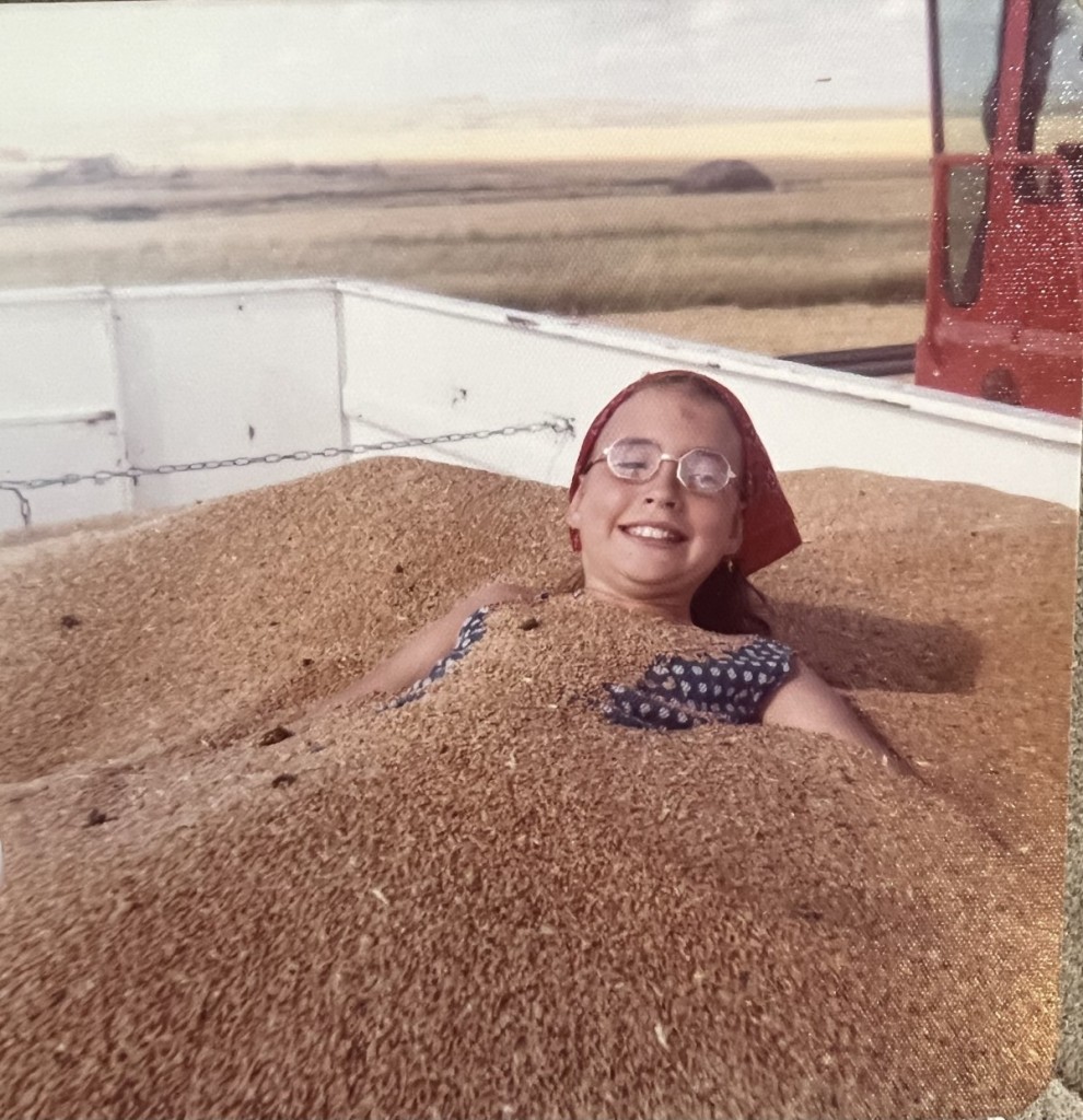 A young girl wearing glasses and a red bandana buried in wheat inside a grain truck, smiling with joy during harvest.