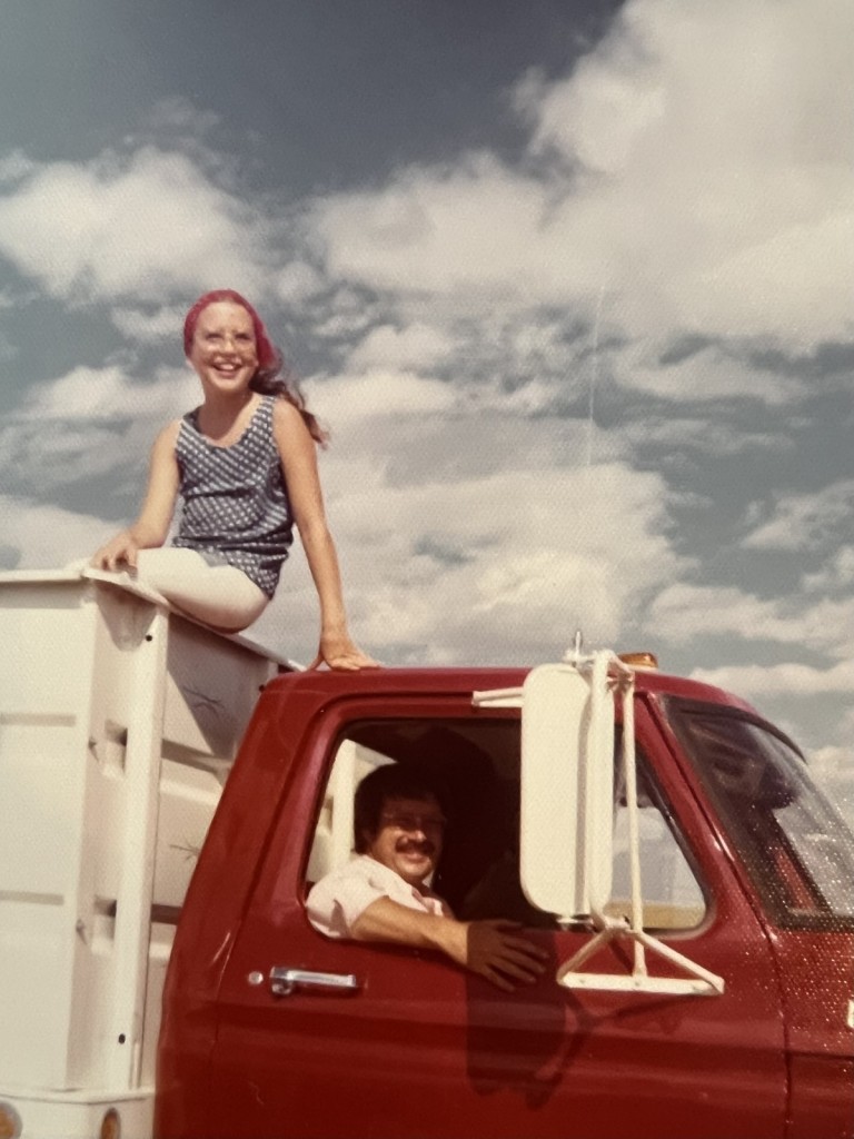 A young girl sitting on top of a grain truck with her dad in the drivers seat, surrounded by combines and harvest fields