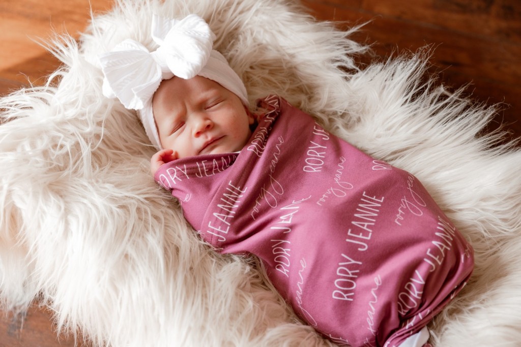 Newborn baby girl wrapped in custom Rory Jeanne swaddle on white faux fur blanket