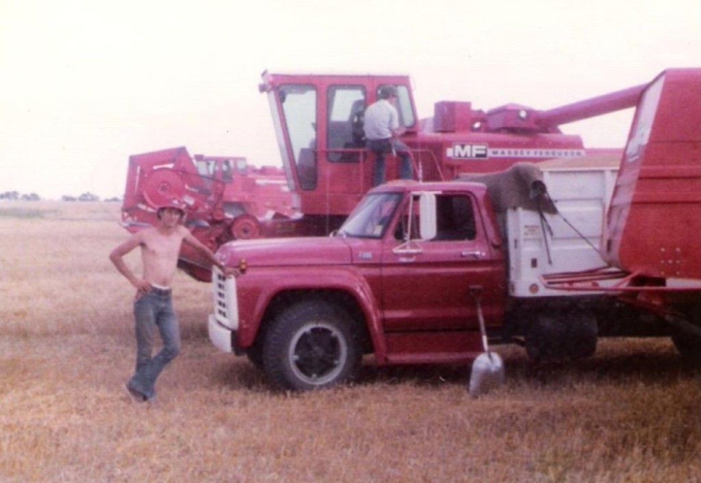 A vintage photo of a shirtless young man leaning on a red grain truck during wheat harvest, comines in the background.