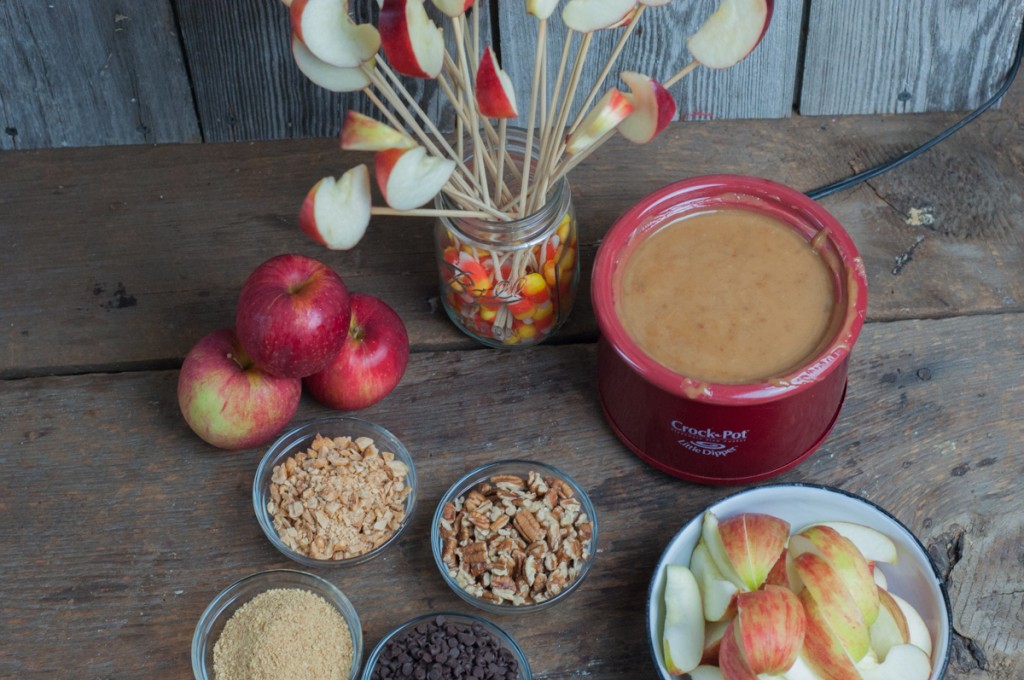 Overhead view of caramel apple dip in a red crockpot surrounded by apples and small bowls of toppings.